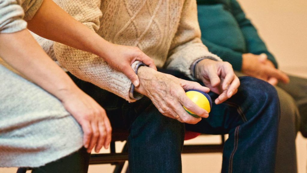 Eine ältere Person erhält Unterstützung von einer Pflegekraft, die im Haus die Hände hält und Mitgefühl zeigt.